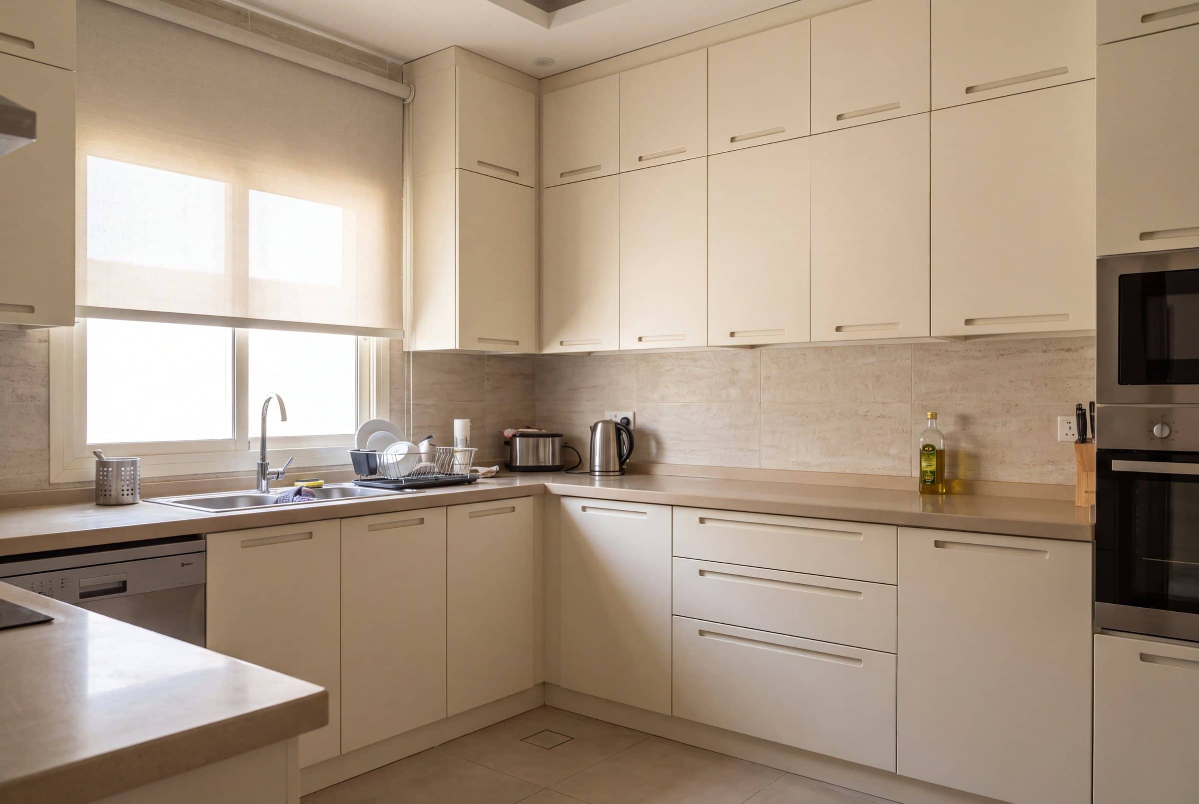 Apartment kitchen with warm-white lacquered doors and routed pull grooves, a beige worktop and a window over the sink